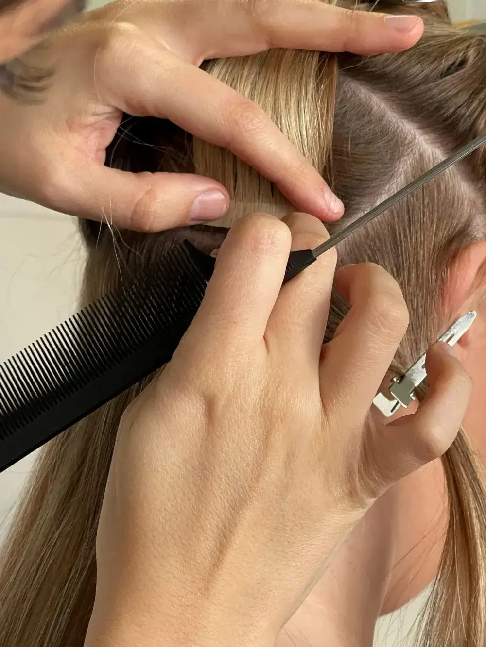 Gros plan sur une main qui prépare une section de cheveux blonds à l’aide d’un peigne à queue avant la pose d’extensions adhésive